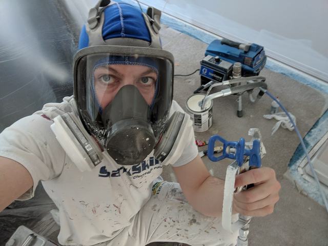 Justin in a full-face respirator holding an airless spray gun next to a Graco sprayer